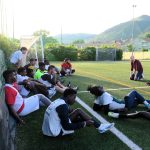 L'allenatore di Casa Sport parla coi i suoi giocatori sul terreno di gioco, tutti seduti accanto alla porta a bordocampo